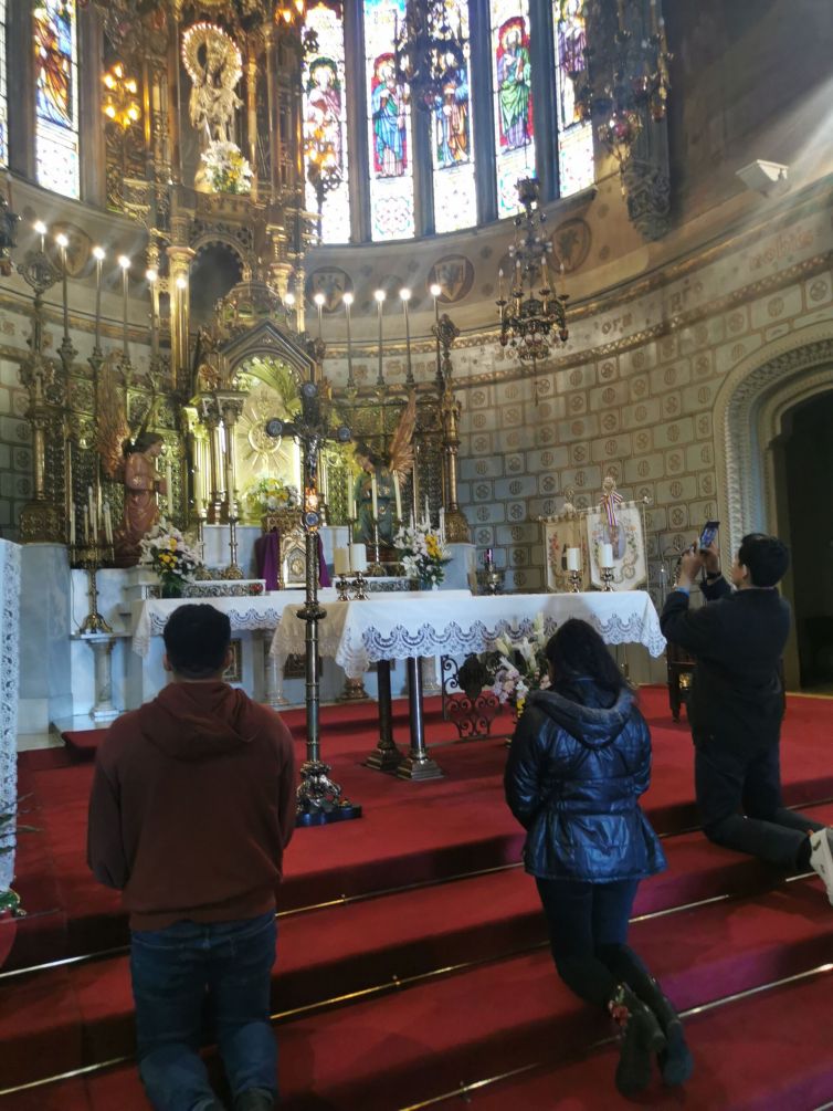 Dos personas de rodilla ante el altar del Santuario rezan a San José de la Montaña y otra persona, también de rodillas, hace una foto a la imagen.