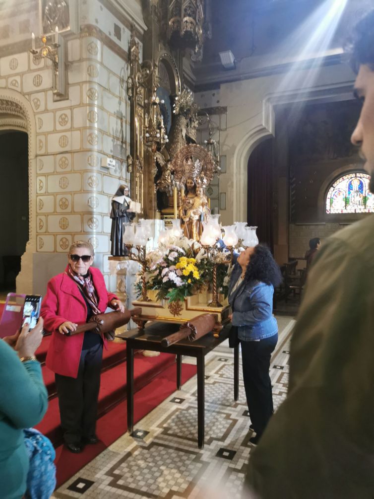 Señora con abrigo rosa posa junto a la imagen de San José de la Montaña mientras le hacen una foto. A la derecha otra devota toca la imagen y en el recuadro derecho se ve el inicio de la cola para acercarse.